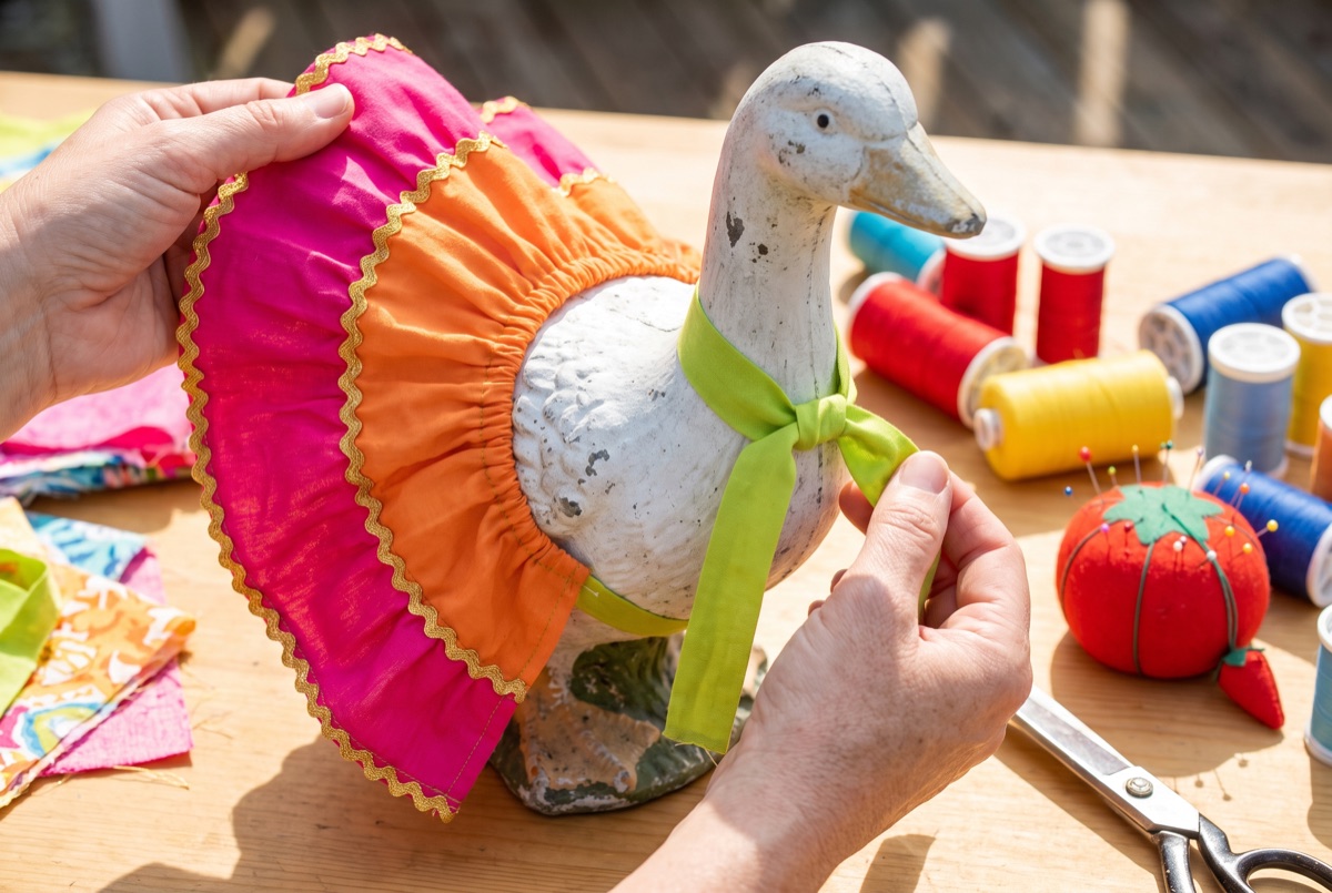 Two hands fastening a hot pink and orange ruffled skirt onto a weathered concrete goose, with spools of colourful thread, pins and a tomato pincushion scattered on a sunlit wood table.
